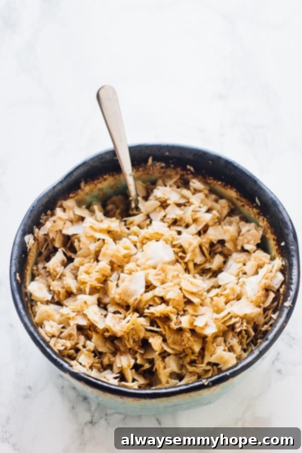 These gluten-free vegan bacon crumbles are made out of coconut, and taste just like real bacon. They're salty, smoky, and sweet! A bowl of unsweetened coconut flakes, beautifully coated in a dark, smoky, and sweet marinade, with a spoon resting inside, ready for baking.