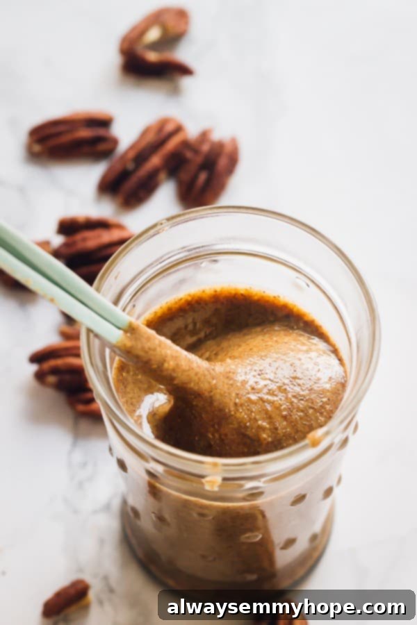 Creamy Homemade Pecan Butter Scoop A spoon elegantly scooping creamy homemade pecan butter from a transparent glass jar, highlighting its smooth texture.