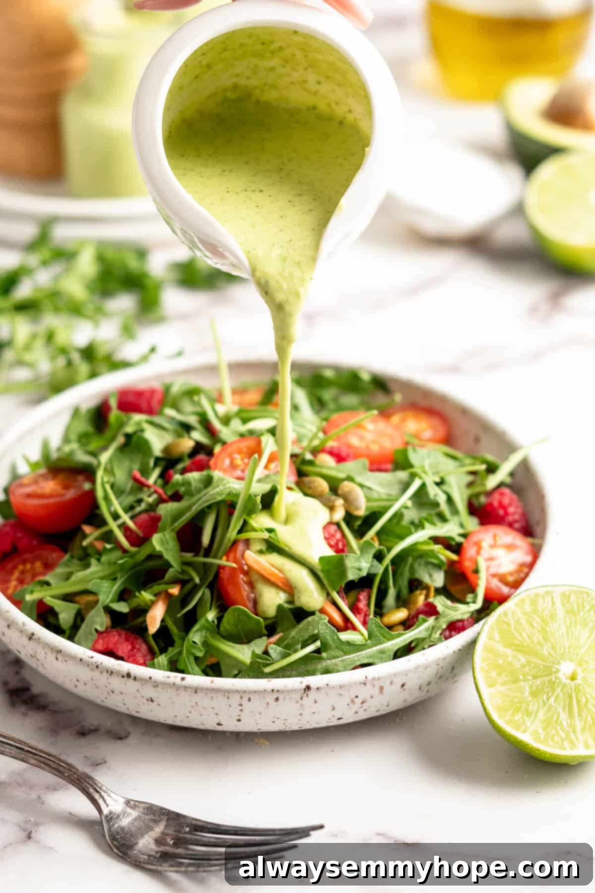 Naturally Creamy Vegan Avocado Dressing 6 A hand pouring generous amount of vibrant green creamy vegan avocado dressing from a small pitcher onto a colorful fresh salad in a bowl.