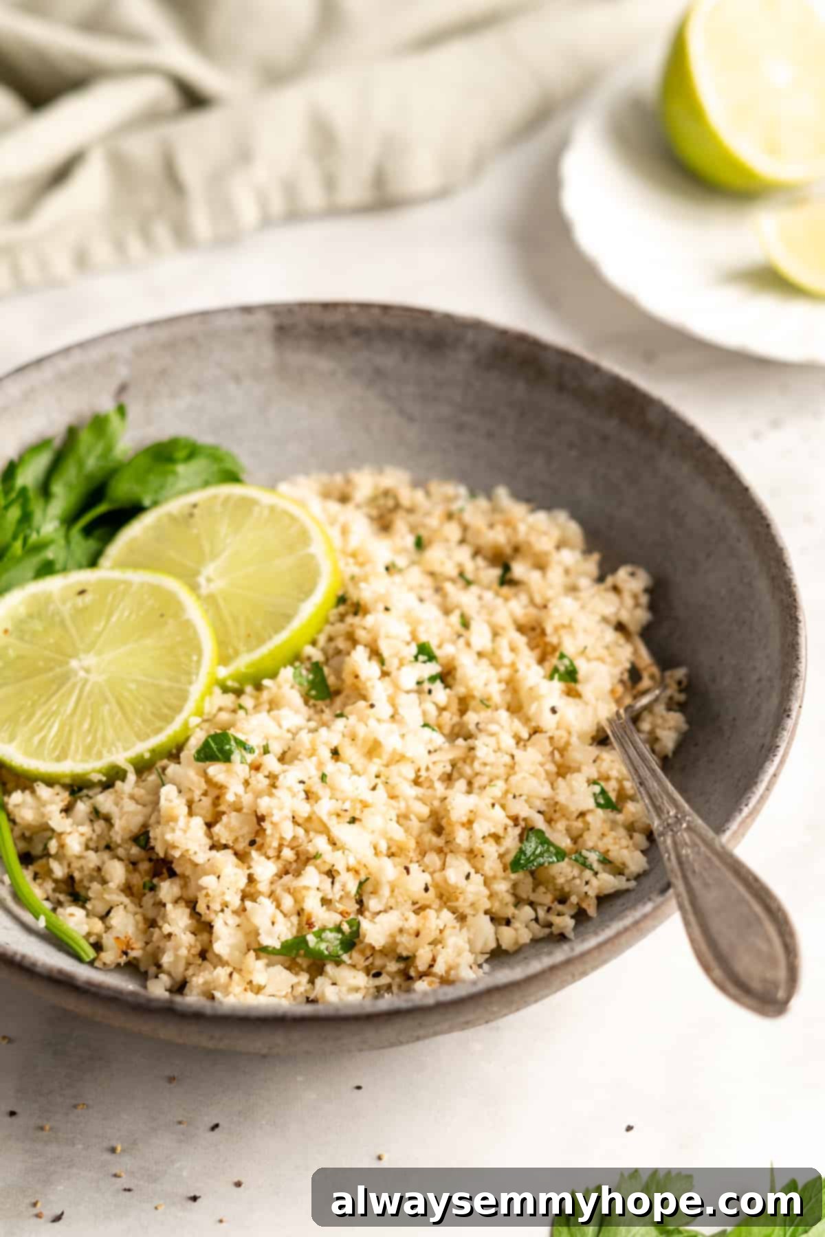 A bowl of cooked cauliflower rice with a fork, garnished with fresh lime slices and parsley, ready to be enjoyed.