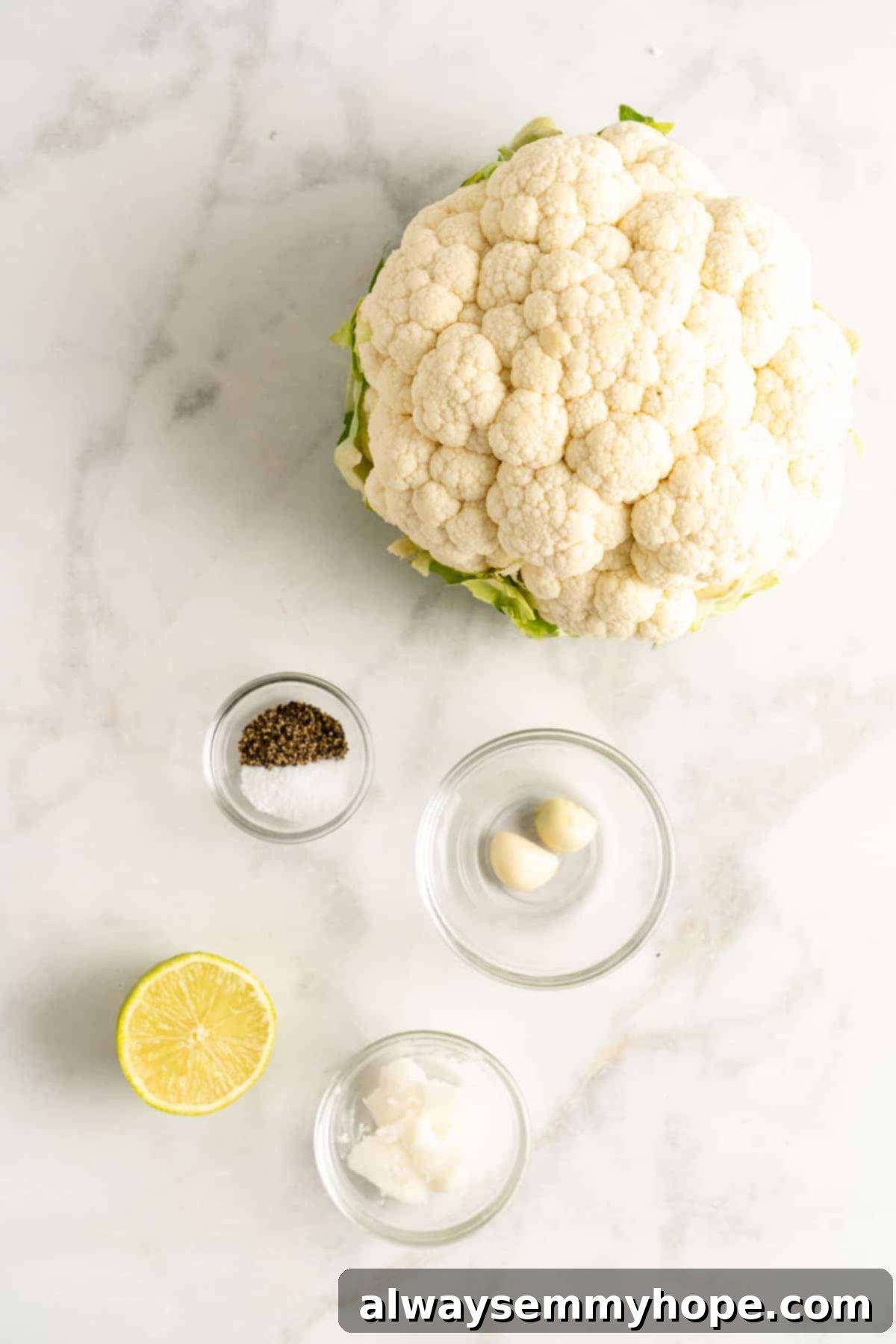 Overhead shot of fresh cauliflower rice ingredients laid out: a head of cauliflower, garlic cloves, a lime, and a bottle of coconut oil.