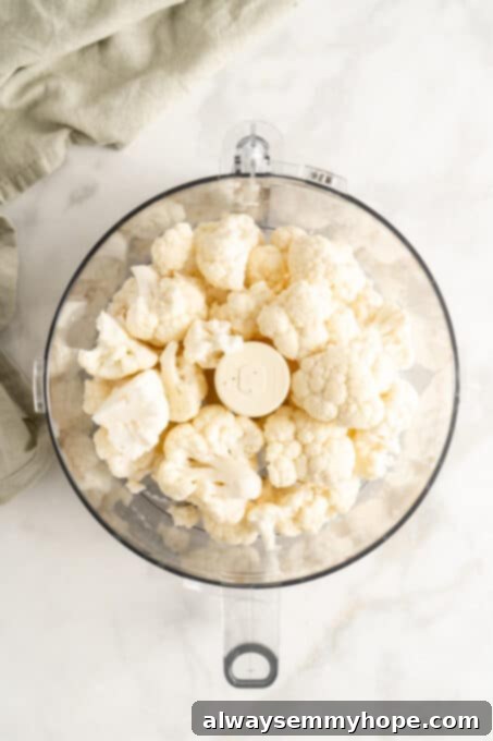 Close-up overhead view of cauliflower florets inside a food processor bowl, ready to be pulsed.