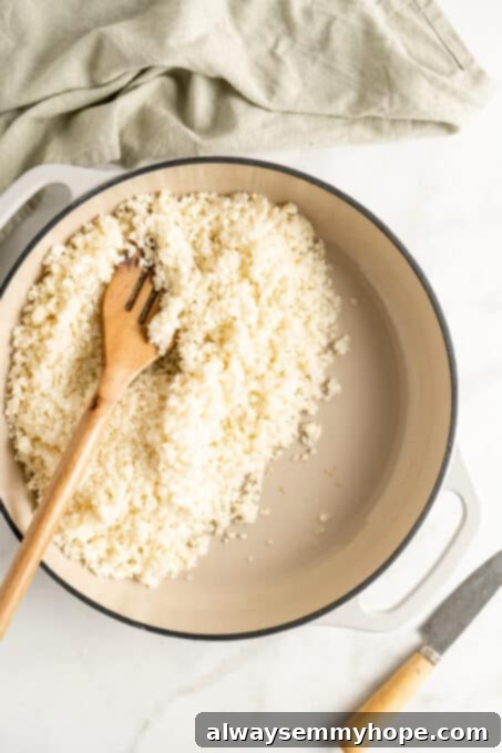 Overhead view of uncooked cauliflower rice in a white cast iron skillet, stirred with a wooden spatula.
