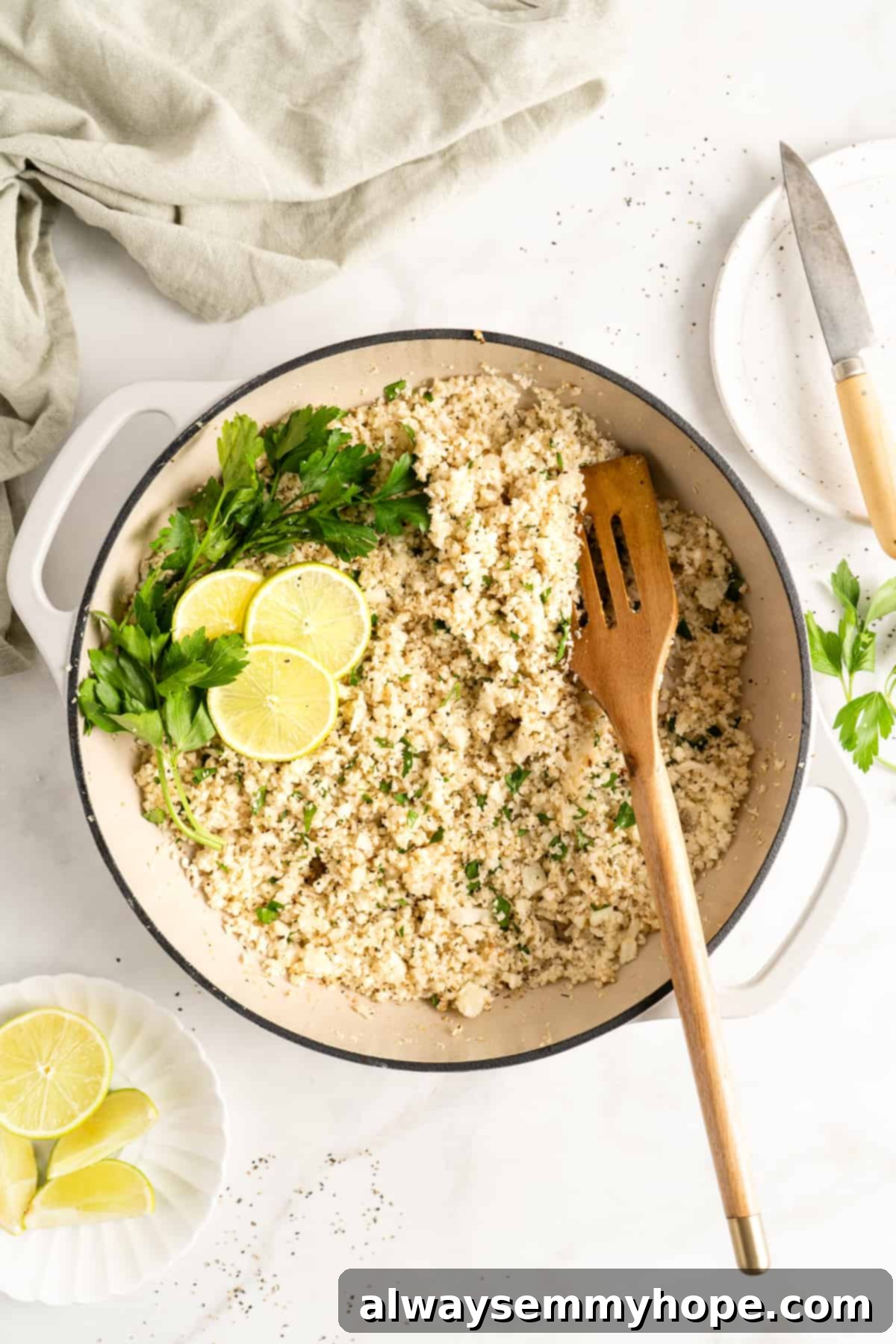 Overhead view of cooked cauliflower rice in a white cast iron skillet with a wooden spatula, elegantly garnished with lime wedges and fresh parsley.