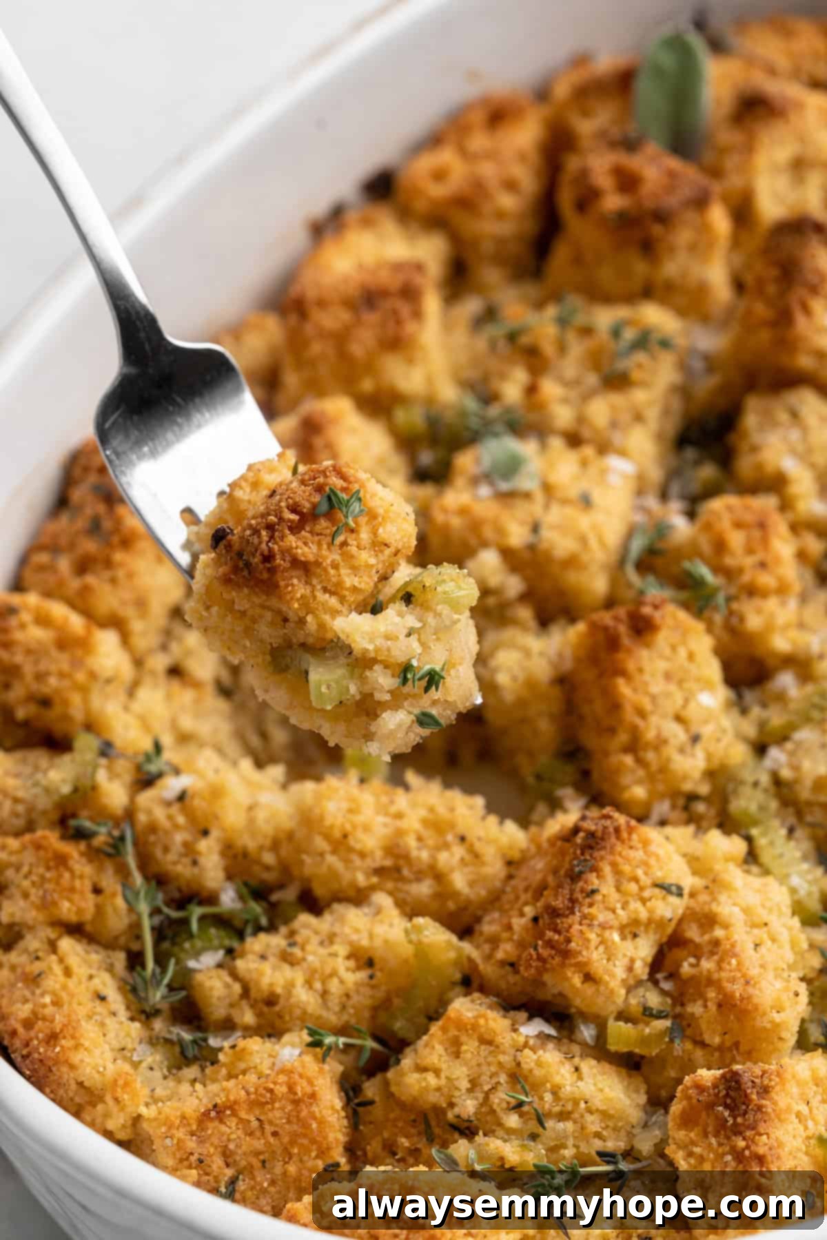 A fork holding a perfect bite of warm cornbread stuffing, lifted above the baking dish, garnished with fresh herbs