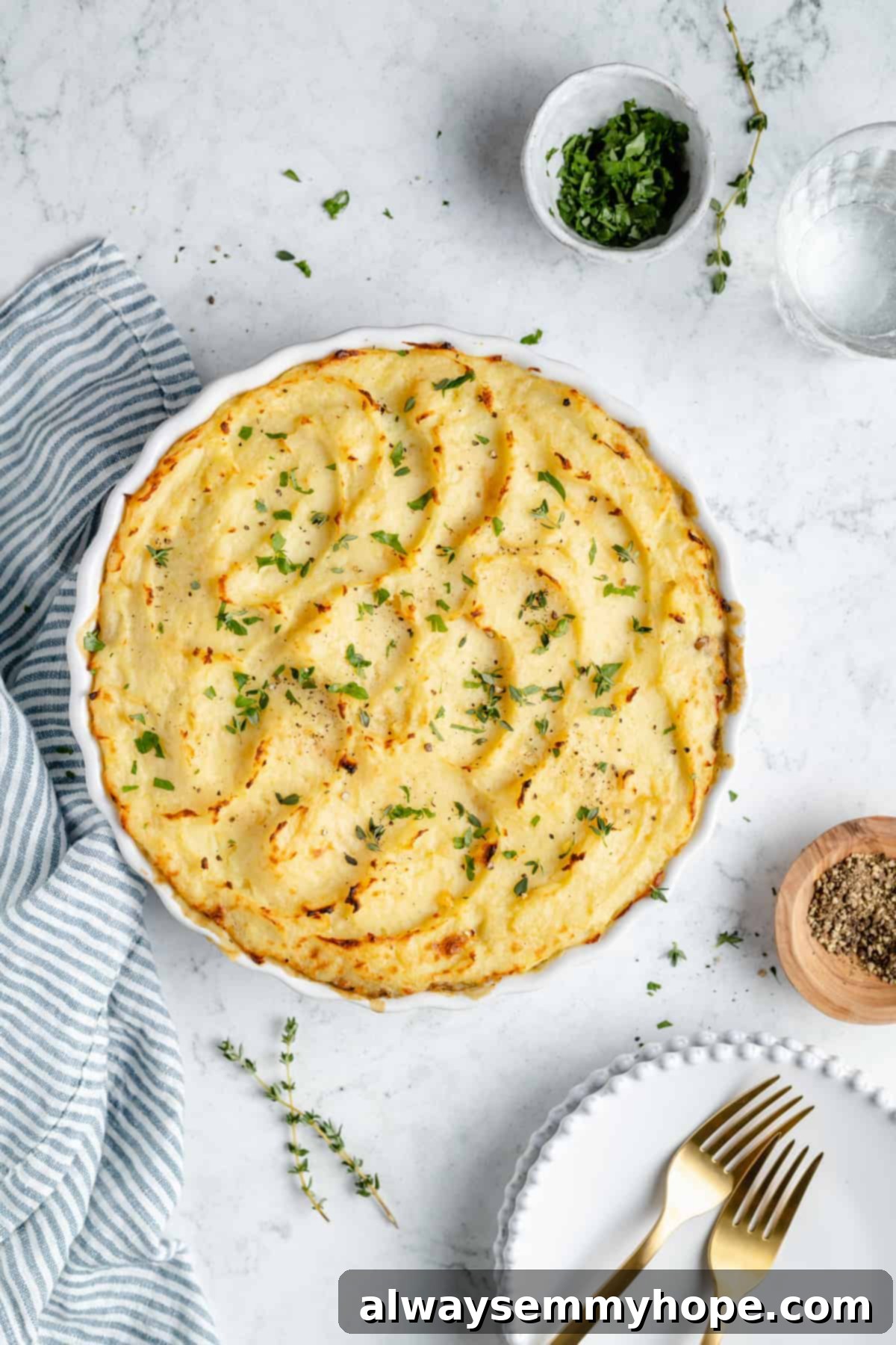 Hearty Vegan Shepherd's Pie 12 Overhead view of a baked Vegan Shepherd's Pie in a serving dish, with plates, forks, and fresh herb garnishes arranged around it