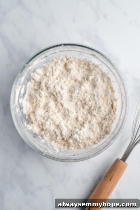 A glass mixing bowl containing the combined dry ingredients for vegan biscuits, showing a coarse, sandy texture.