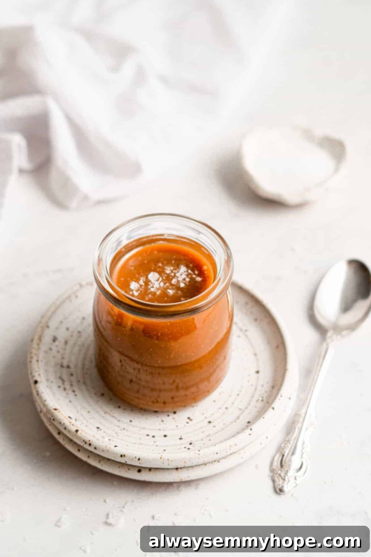 Simple Plant-Based Caramel 8 A jar of freshly made vegan caramel sauce, with a spoon beside it, on a white plate.