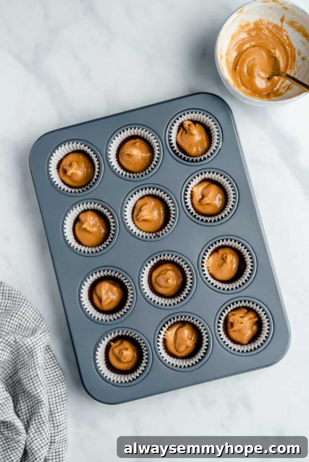 Melted chocolate base and dollops of peanut butter filling layered in mini muffin liners within a muffin tin, mid-assembly.