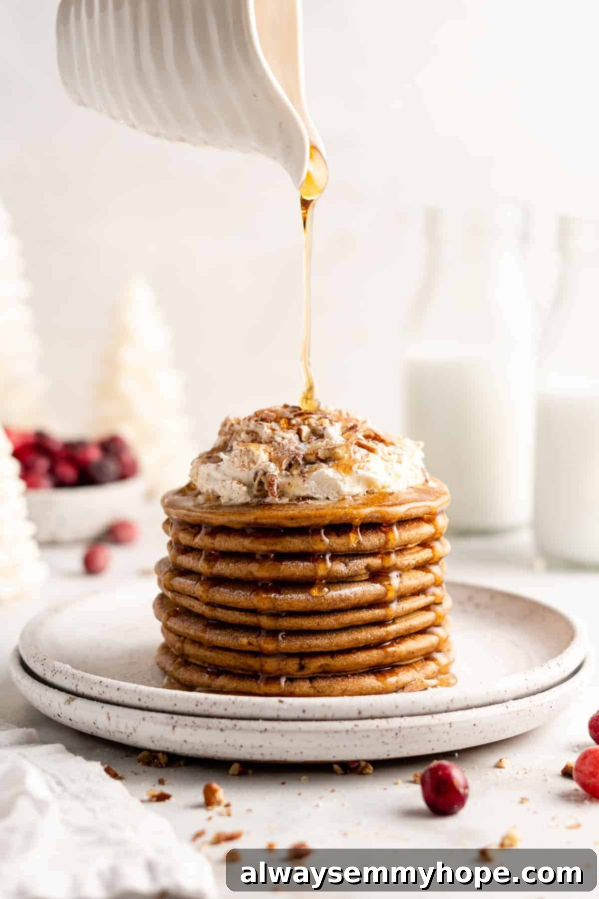 Pouring maple syrup over a stack of fluffy vegan gingerbread pancakes topped with coconut whipped cream and chopped nuts, capturing the perfect plant-based Christmas breakfast.