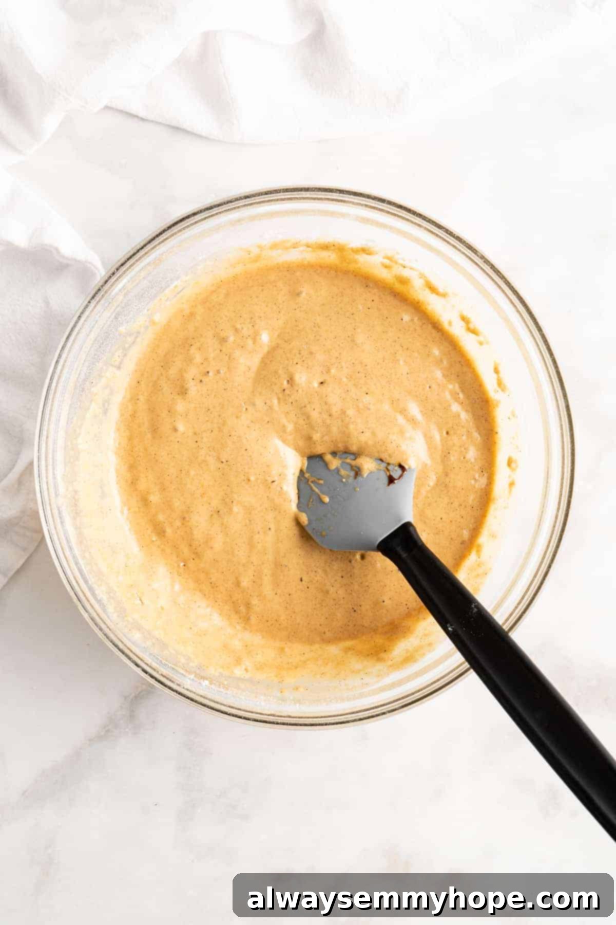 A plastic spoon rests in a glass mixing bowl filled with freshly prepared, slightly lumpy vegan gingerbread pancake batter, ready for cooking.