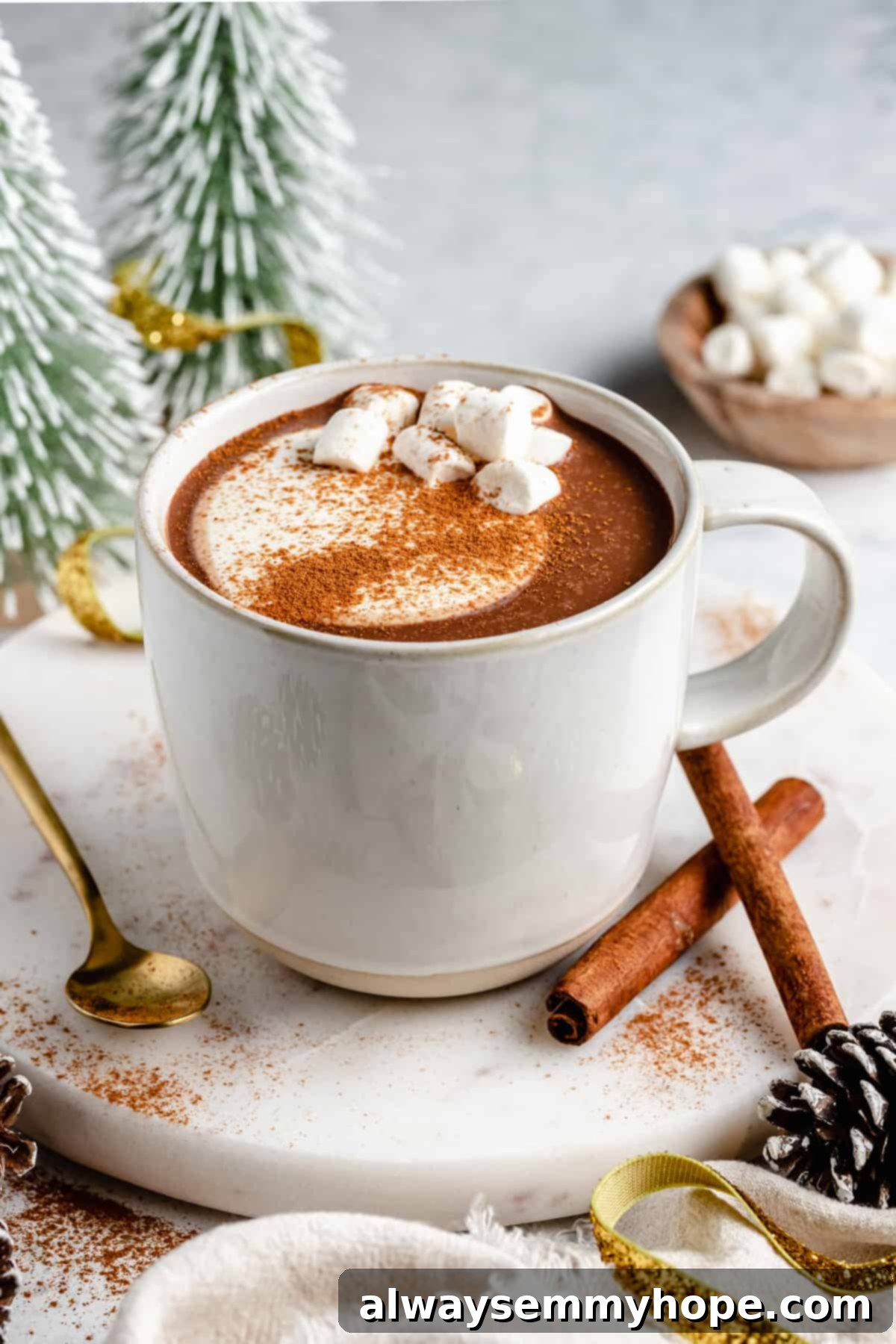 A cozy mug of vegan hot chocolate, garnished with a cinnamon stick, sits on a round marble slab with a spoon nearby.