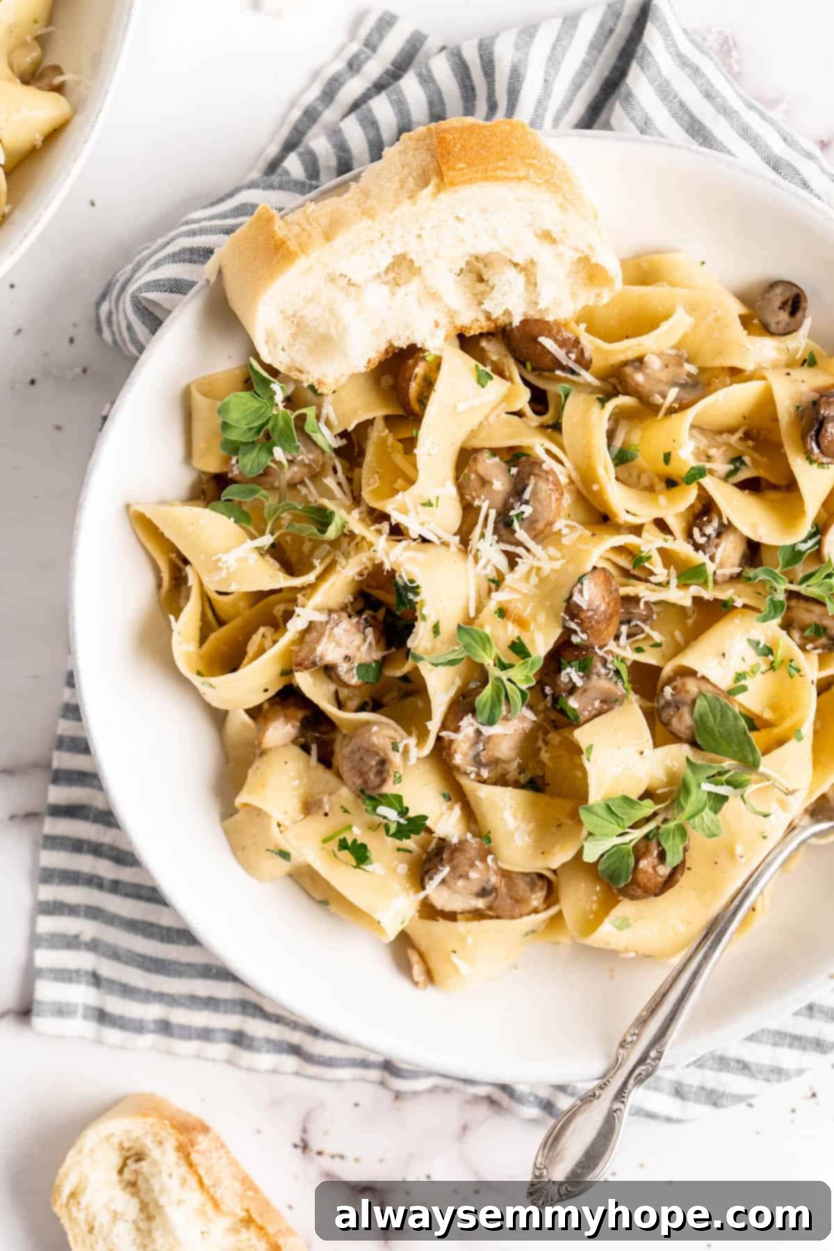 Overhead view of bowl of Garlic Mushroom Pasta with slice of baguette, highlighting its creamy texture and vibrant garnishes.