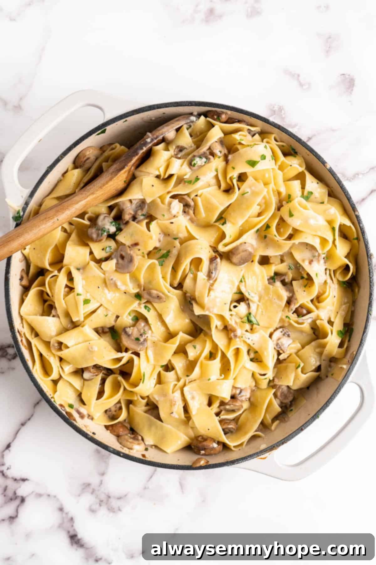 Overhead view of Garlic Mushroom Pasta in an enamel pan with a wooden spoon, showing the pasta being tossed with the sauce.