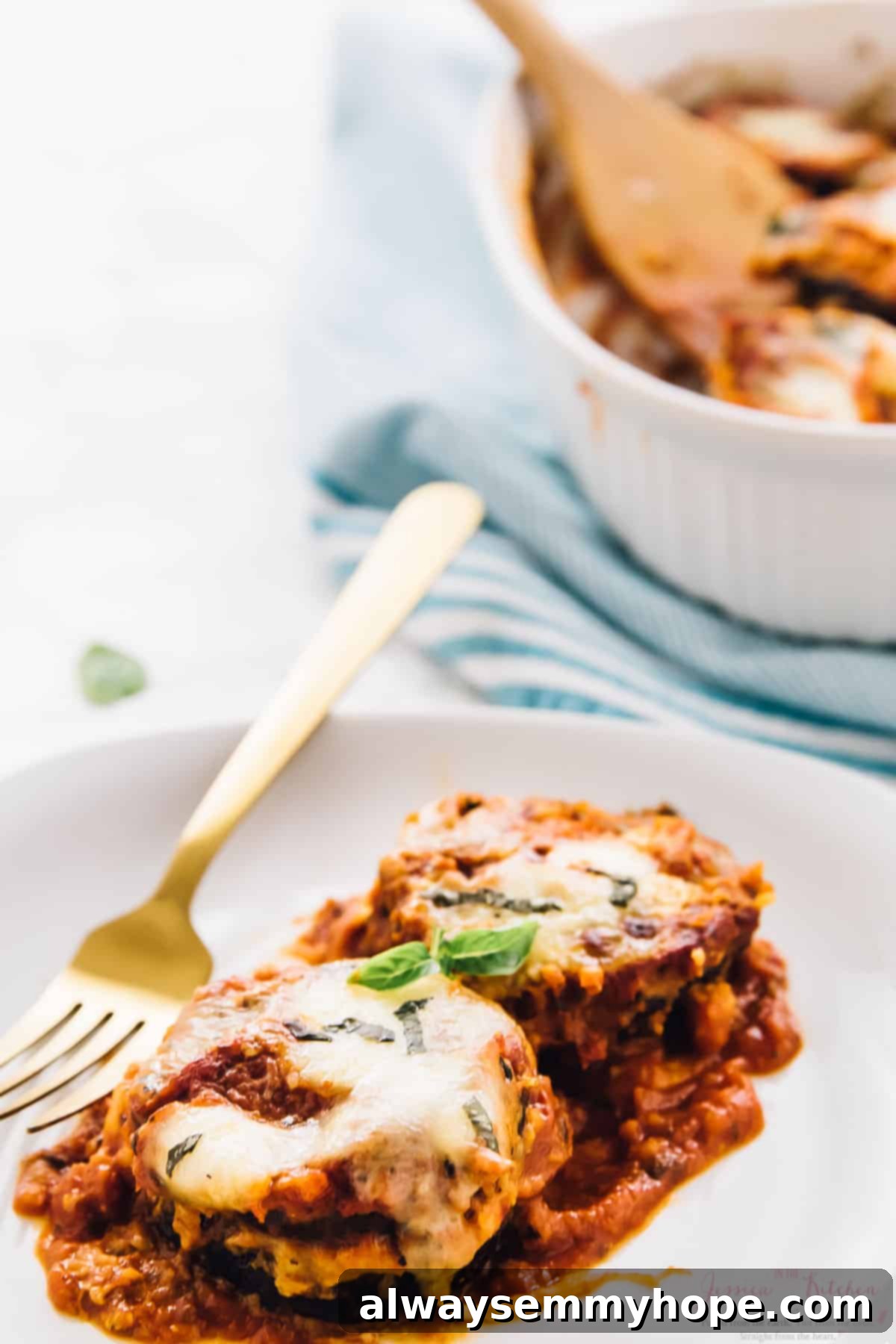 A slice of Baked Eggplant Parmesan on a white plate with a gold fork, garnished with fresh basil.