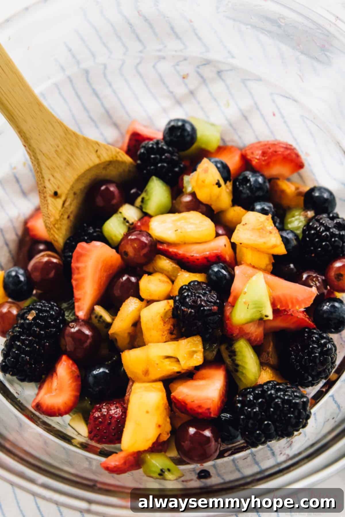 Vibrant Fruit Medley with Zesty Maple-Lime Drizzle 6 A close-up shot of a wooden spoon gently stirring an abundant mixture of colorful fruits in a clear glass bowl, showcasing the textures and fresh colors.