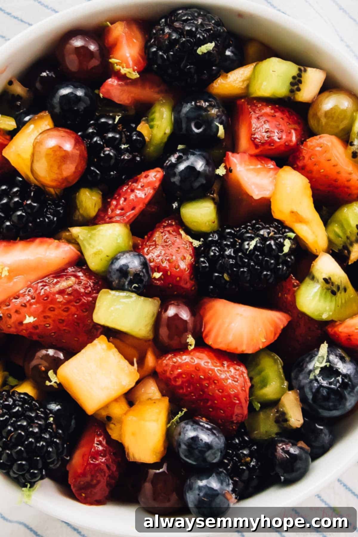 Vibrant Fruit Medley with Zesty Maple-Lime Drizzle 7 Overhead view of a vibrant rainbow fruit salad in a white bowl, elegantly placed on a light-colored striped cloth, highlighting its freshness.