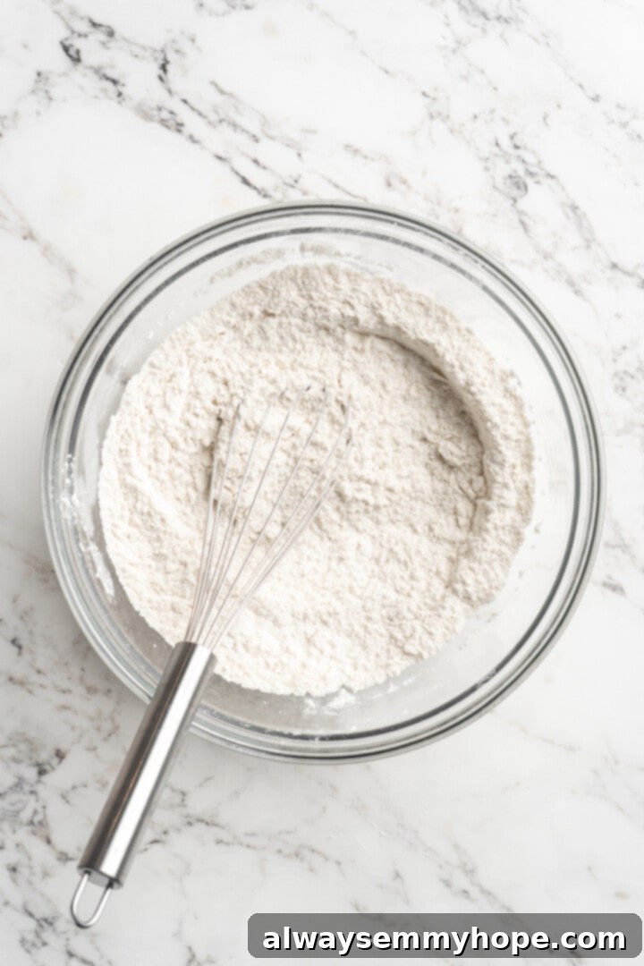 Overhead view of dry ingredients (flour, baking powder, baking soda, salt) in a bowl with a whisk, ready to be combined.