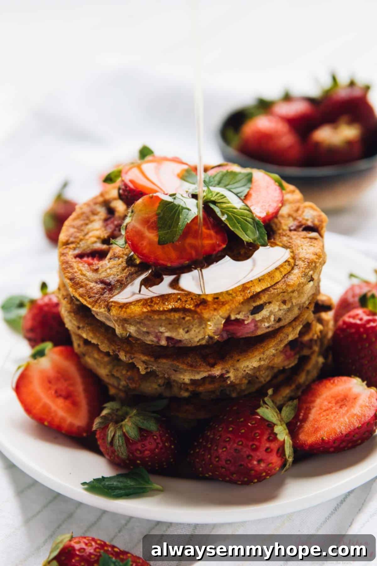 Drizzling maple syrup onto a tall stack of perfectly golden vegan gluten-free strawberry pancakes, with fresh strawberries scattered around the plate.