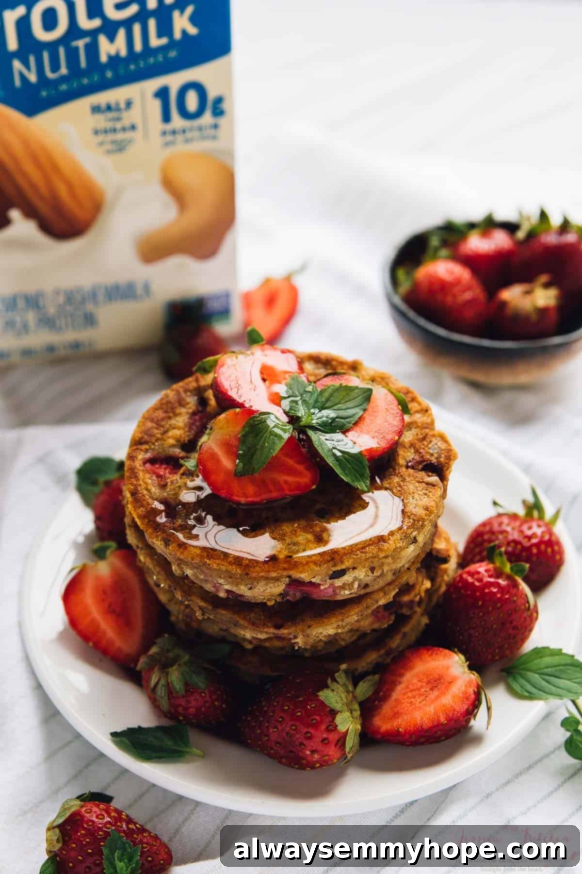 A stack of golden strawberry pancakes adorned with fresh whole strawberries and a generous drizzle of maple syrup on a white plate.