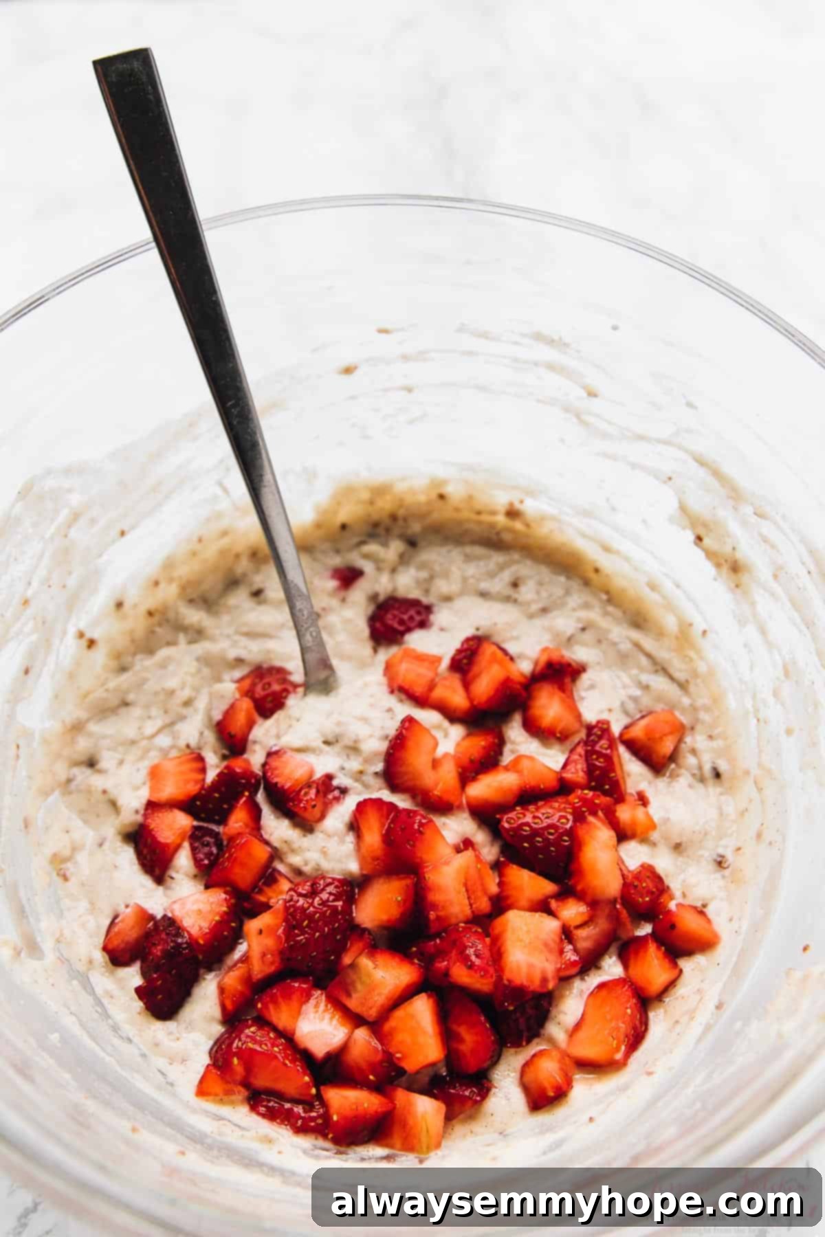 Chopped fresh strawberries being gently stirred into a creamy, thick gluten-free vegan pancake batter in a clear glass mixing bowl.