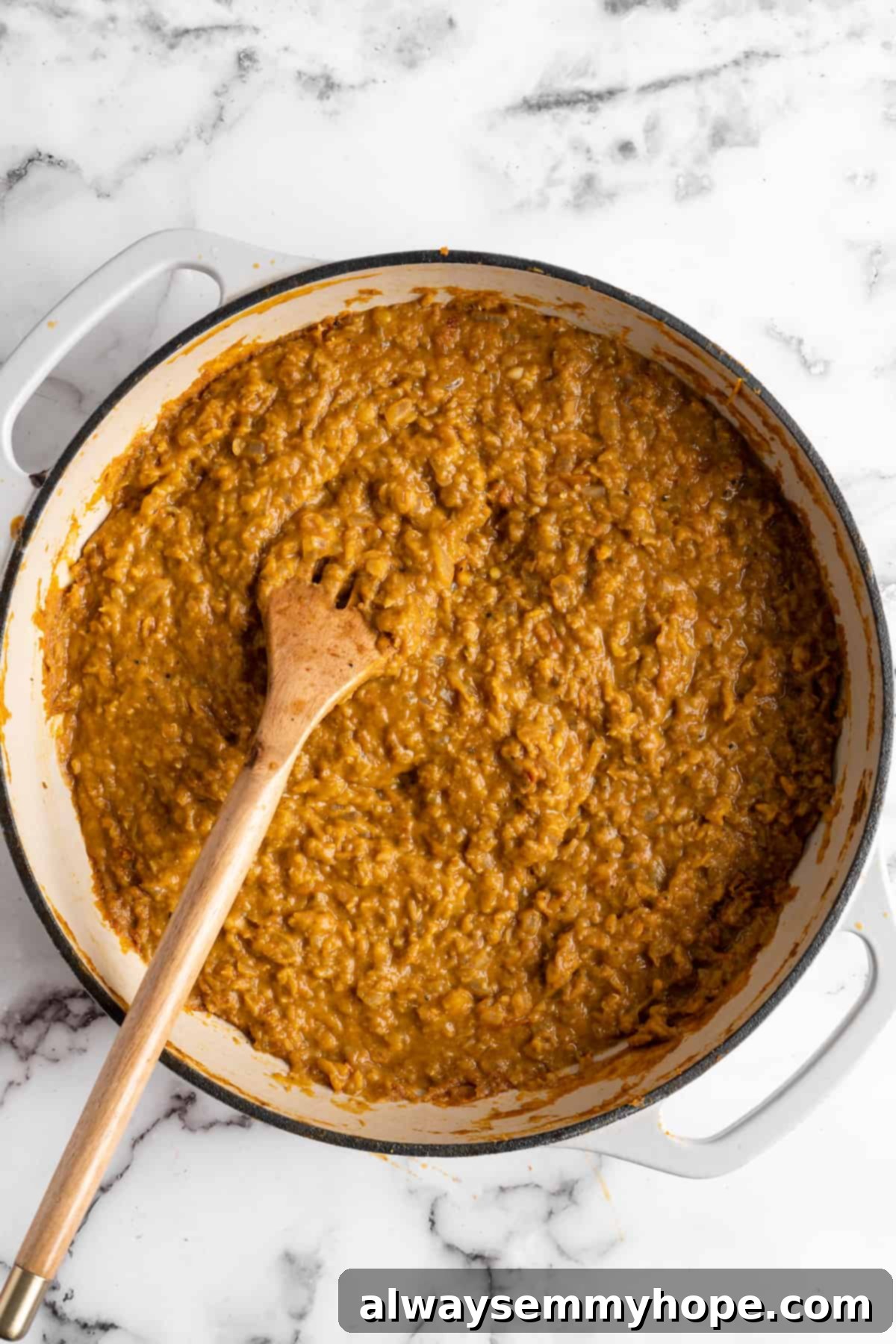 This red lentil curry is classic vegan comfort food! It’s a flavorful one-pot recipe that comes together in under an hour. An inviting overhead shot of finished red lentil curry in a pot, showing its creamy texture, with a wooden spoon resting in it, ready to be served.