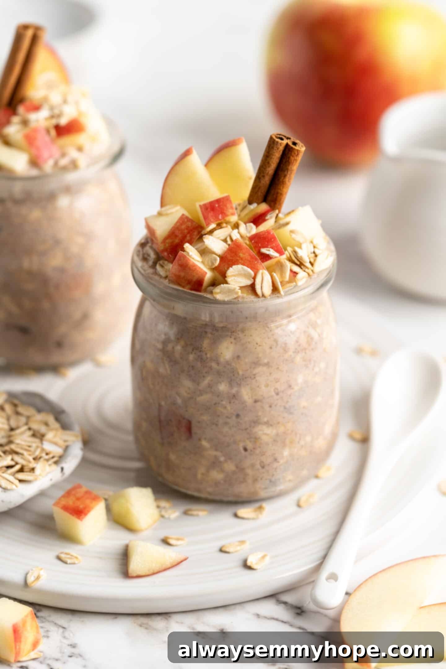 Apple pie overnight oats are packed with crisp apples, warm cinnamon, rich almond milk, and decadent maple syrup, and are an easy breakfast. Two jars of apple pie overnight oats, garnished with cinnamon sticks, rolled oats, and apple slices, with a fresh apple in the background.