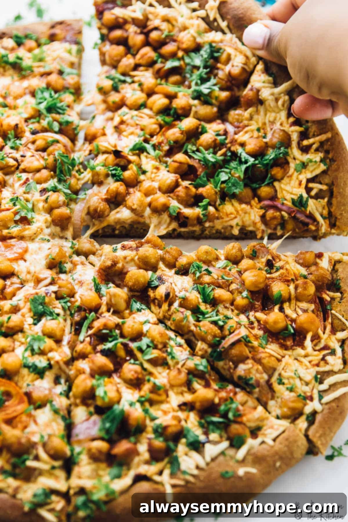 Spicy BBQ chickpea pizza: A delicious vegan alternative to BBQ chicken pizza, packed with flavor and ready in a flash. A hand gently pulling a slice of spicy BBQ chickpea pizza from the whole pie, showing the delicious stretch of vegan cheese and abundant toppings.