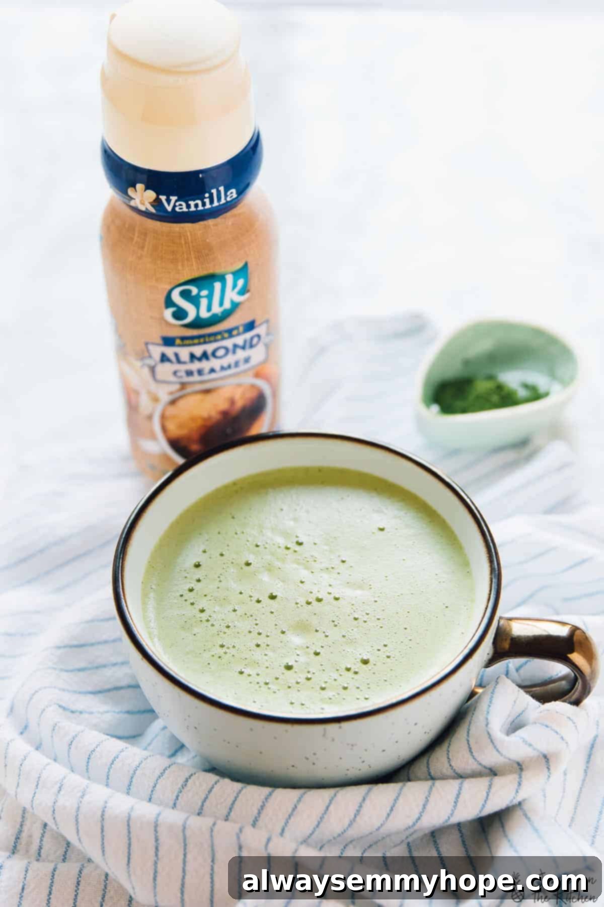 Blender Pumpkin Matcha Latte A Pumpkin Matcha Latte in a pristine white cup, with a complementary bottle of almond creamer blurred in the background, creating a cozy and inviting scene.