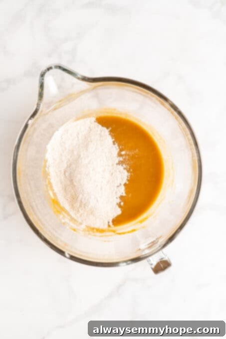 Overhead view of the whisked dry ingredients being gradually added to the wet ingredients (pumpkin, sugar, oil, milk, vanilla) in a glass mixing bowl.
