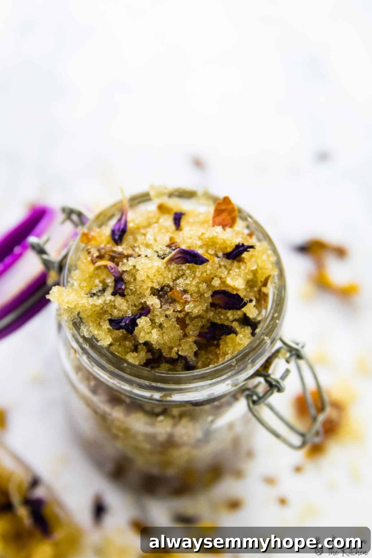A beautifully presented coconut rose sugar scrub in a clear glass jar, with a soft pink hue from the rose petals.
