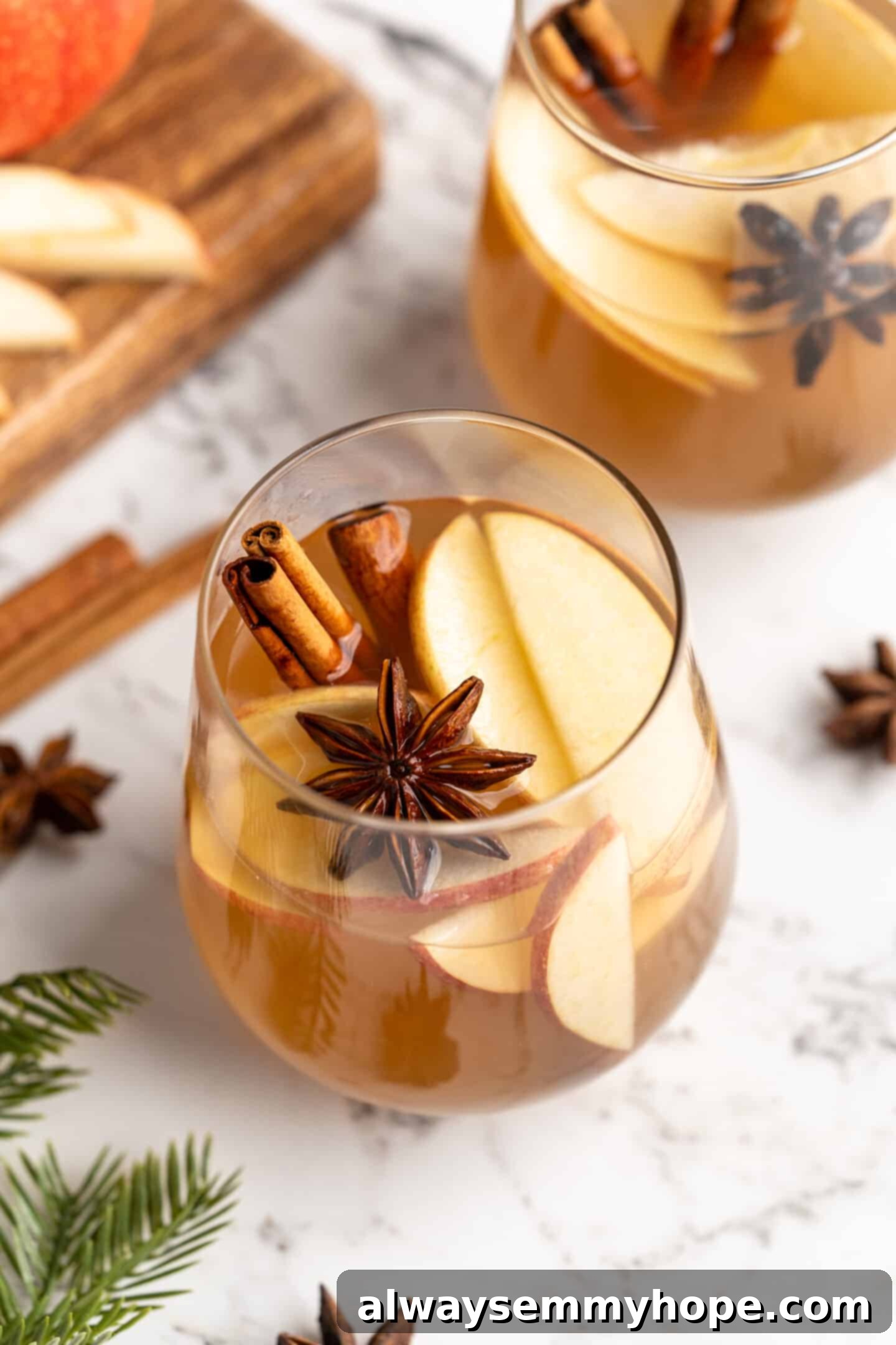 Hot spiced apple cider in a clear glass mug, garnished with whole spices like cinnamon sticks and star anise, and fresh apple slices. The setting evokes warmth and holiday cheer.