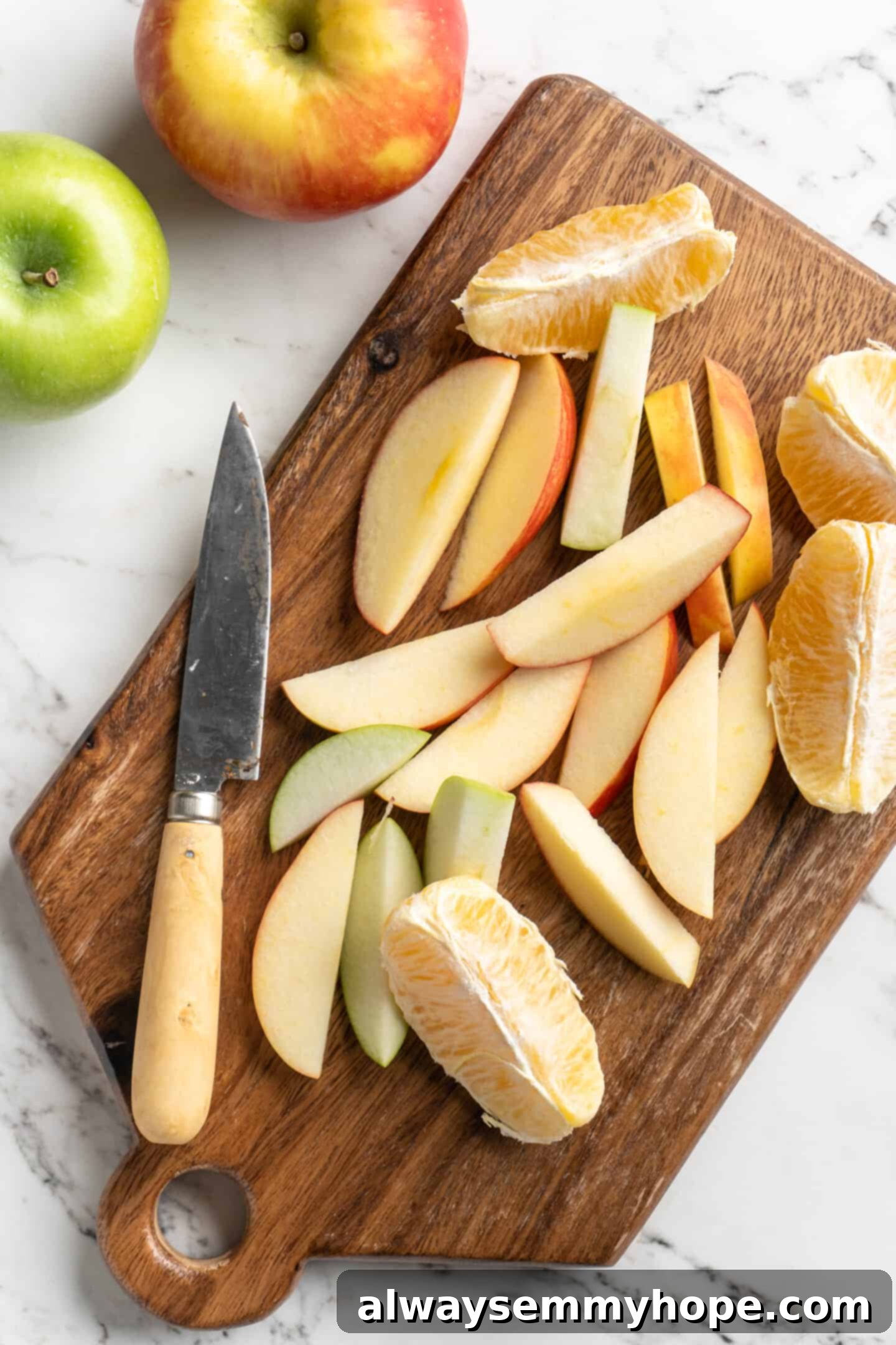 Overhead view of apples and a peeled orange being sliced on a wooden cutting board with a paring knife, ready for the slow cooker.