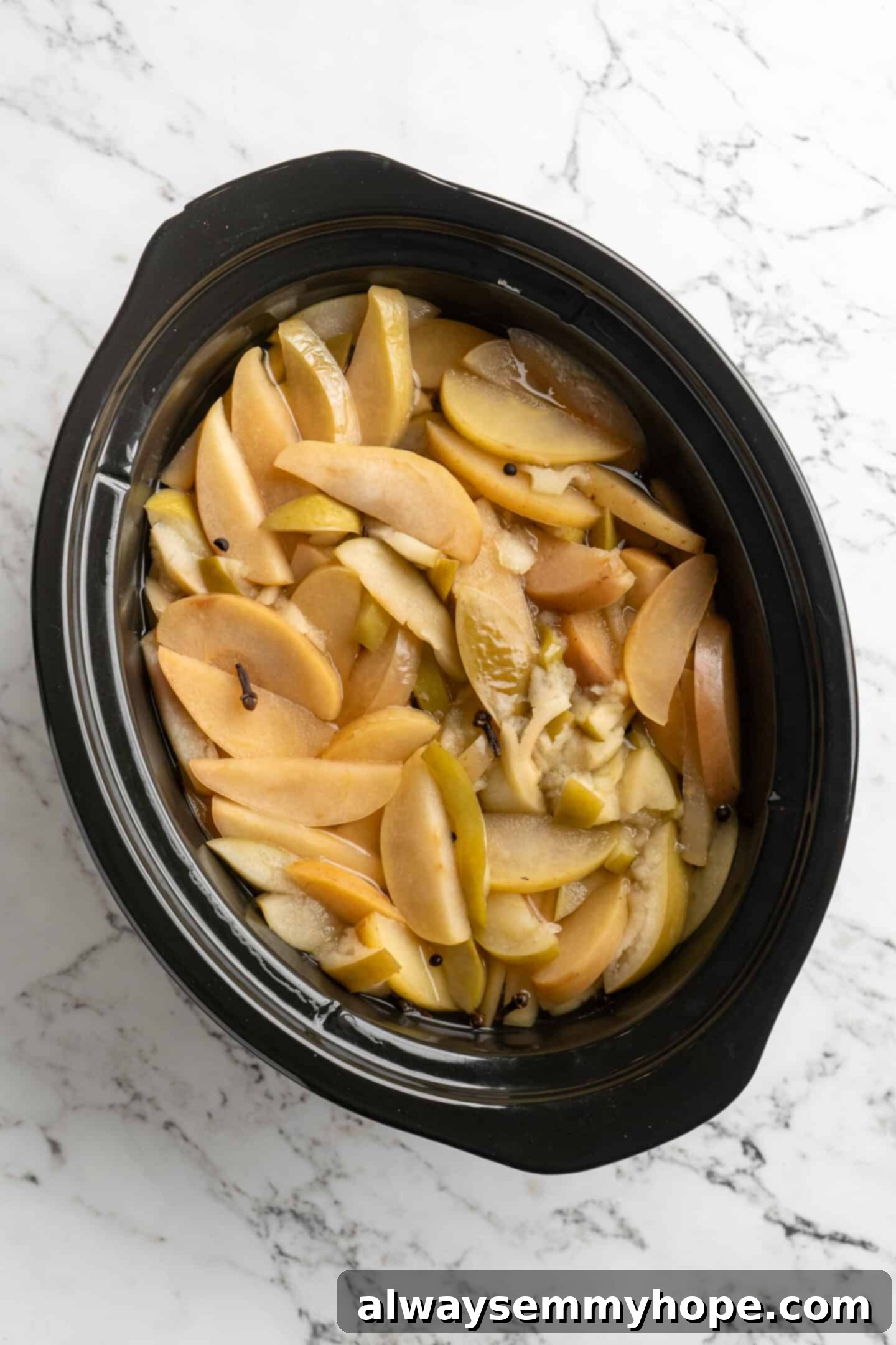 Overhead view of the cooked apples and spices in the slow cooker after several hours, softened and submerged in liquid, ready for mashing.