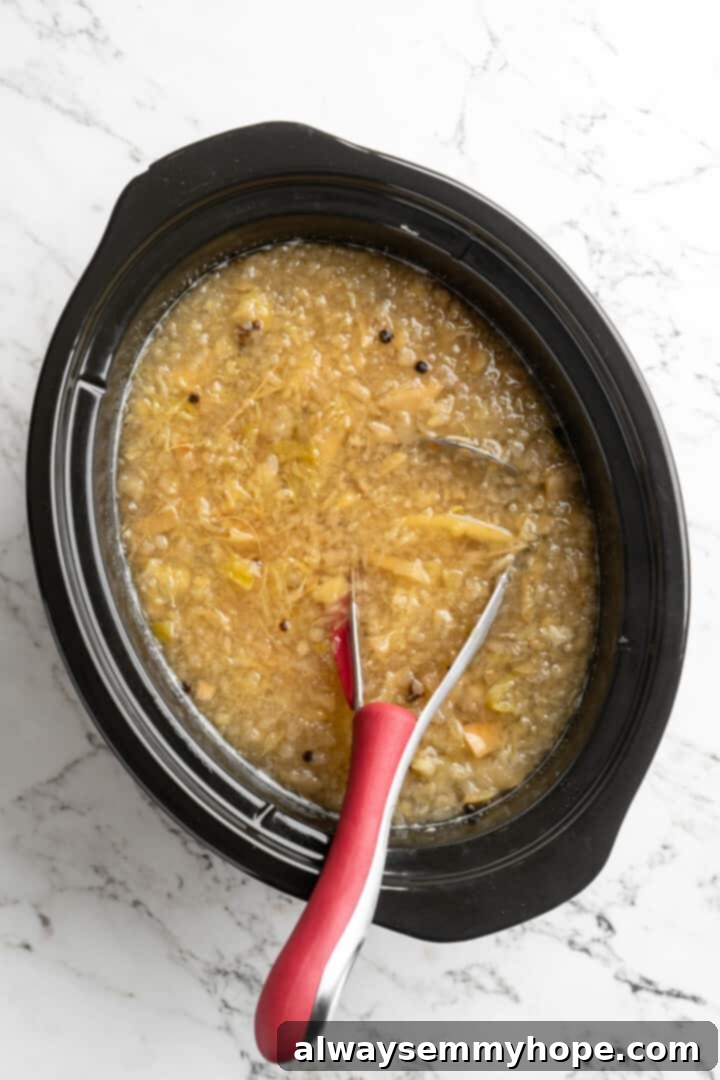 Overhead view of softened apples and oranges being mashed in the slow cooker with a potato masher, releasing their juices and pulp into the cider.