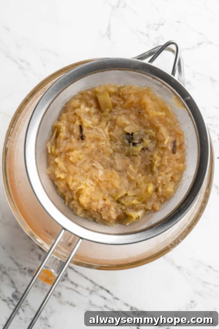 Close-up of the apple cider mixture being poured through a fine mesh sieve into a bowl, separating the liquid cider from the cooked fruit pulp and whole spices.