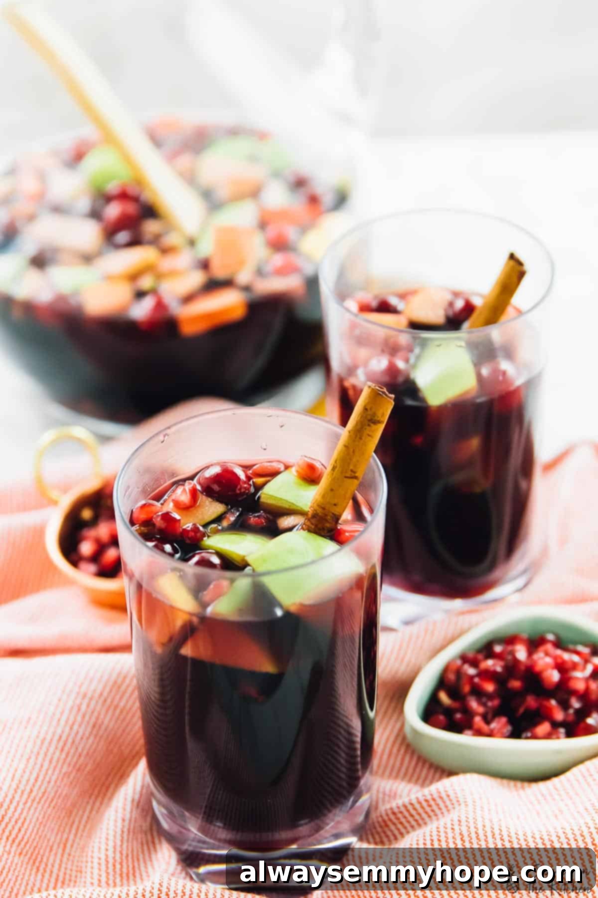 Two glasses of vibrant red sangria, brimming with crisp apple chunks, juicy cranberries, and glistening pomegranate arils, each adorned with a cinnamon stick. Bowls of fresh pomegranate arils sit nearby, with a large pitcher of sangria visible in the soft-focused background, all set for a holiday celebration.