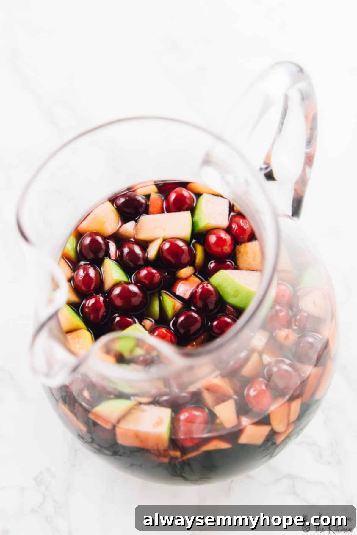 An overhead shot showcasing a elegant glass pitcher filled with homemade sangria. The pitcher is abundantly filled with colorful apple chunks, glistening pomegranate arils, and plump cranberries, reflecting the festive spirit of the drink.