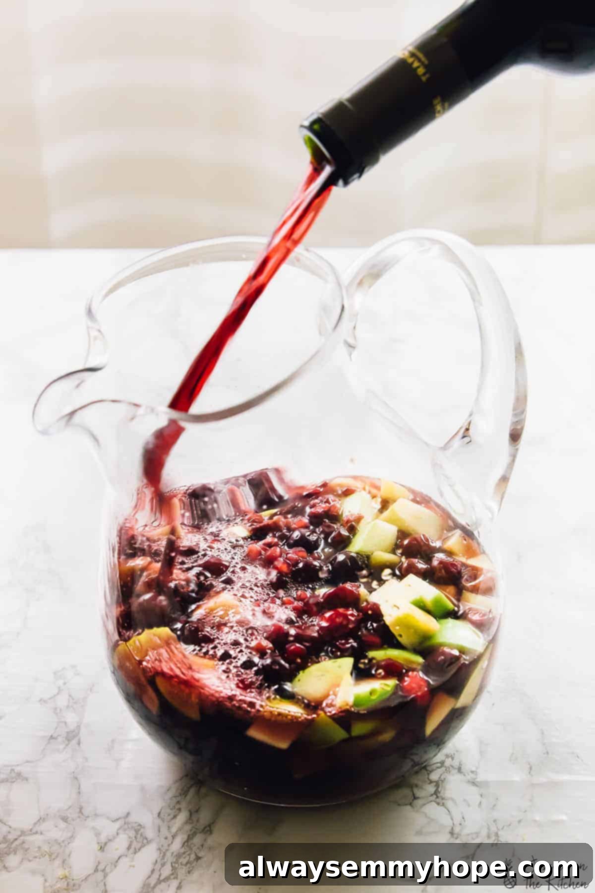 A stream of rich red wine being poured from a bottle into a clear glass pitcher, which is already filled with an enticing mix of vibrant apple pieces, juicy cranberries, and sparkling pomegranate arils, preparing for the perfect sangria.