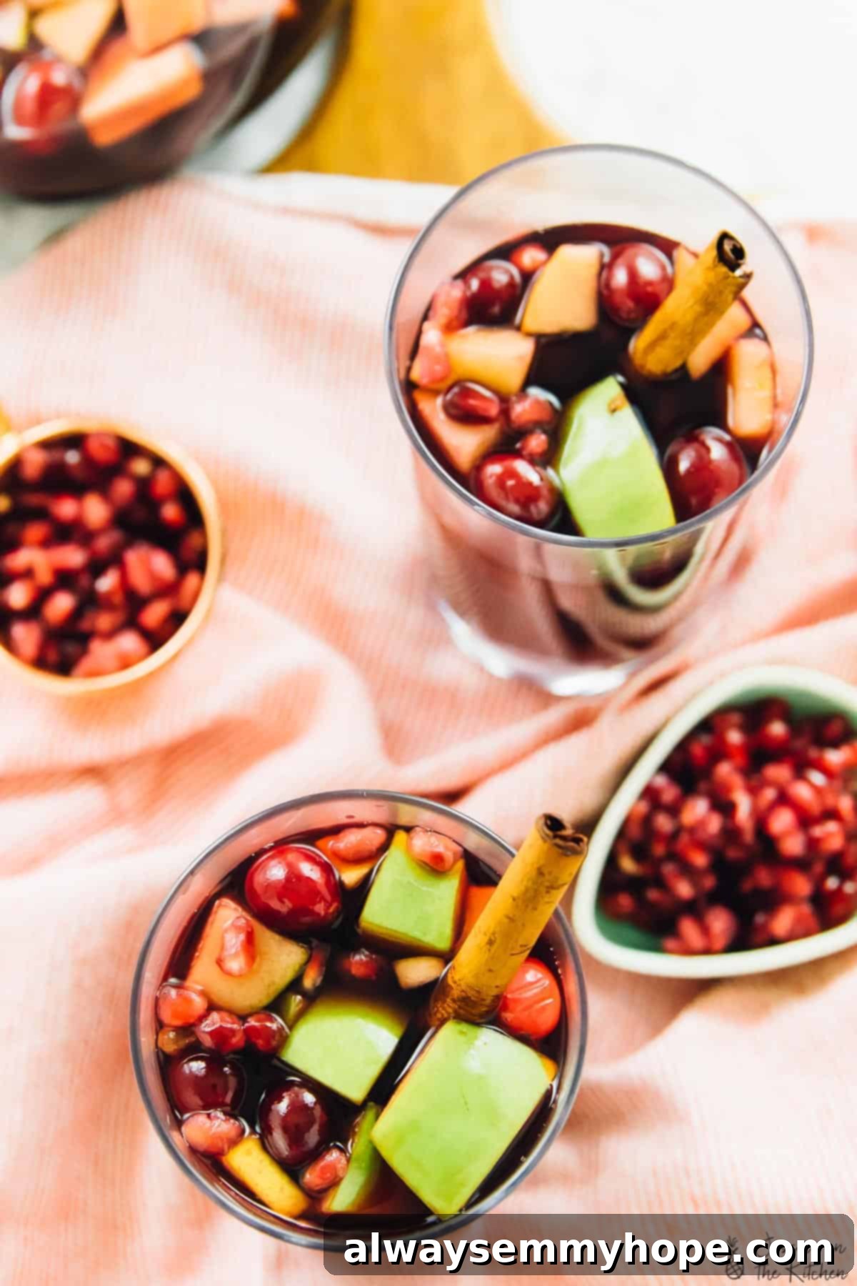 An overhead shot of two beautifully presented glasses of sangria. Each glass is generously filled with crisp apple chunks, juicy cranberries, and sparkling pomegranate arils, with a cinnamon stick elegantly protruding. Two small bowls of additional pomegranate arils sit alongside, completing the festive arrangement.