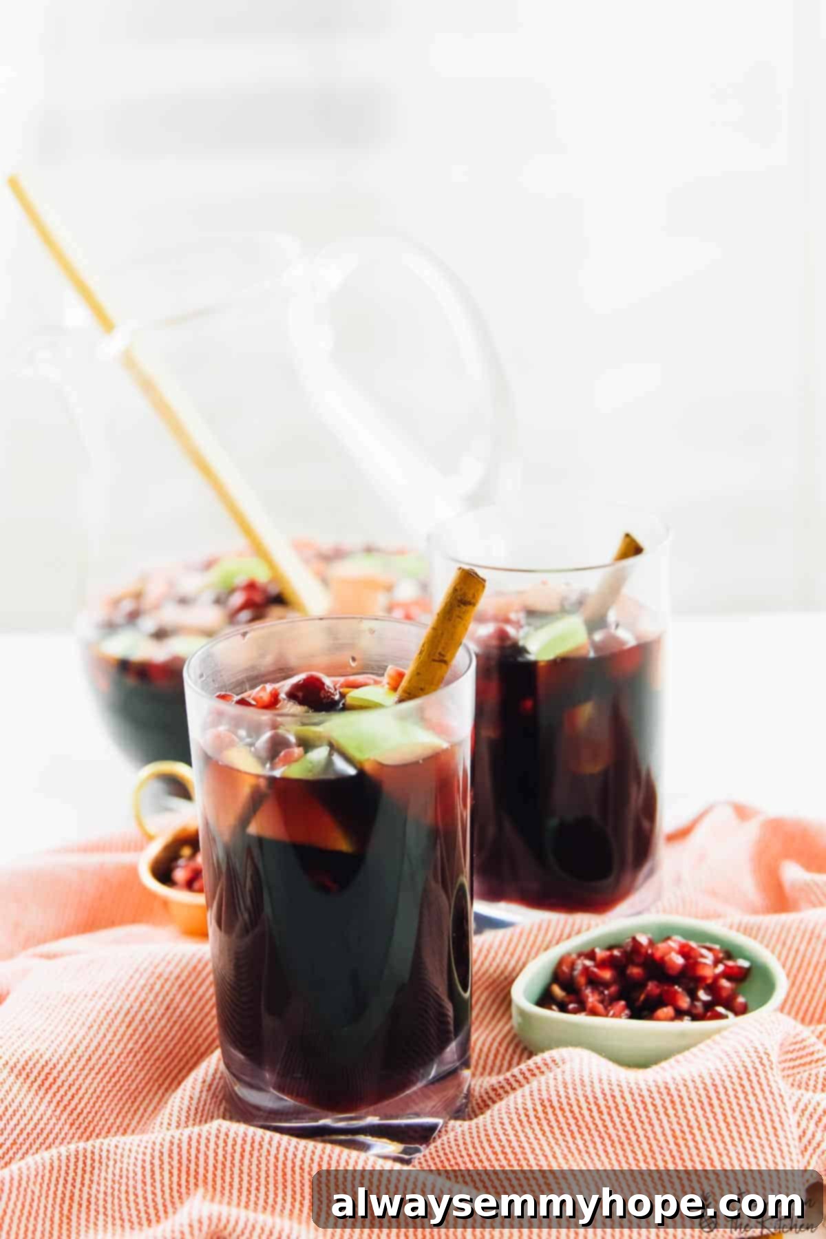 A side view of two elegant, tall glasses of sangria, each garnished with a cinnamon stick and brimming with a colorful medley of apple chunks, vibrant cranberries, and sparkling pomegranate arils. A small bowl of additional pomegranate arils is positioned alongside, with a large pitcher of sangria visible in the background, ready for serving.