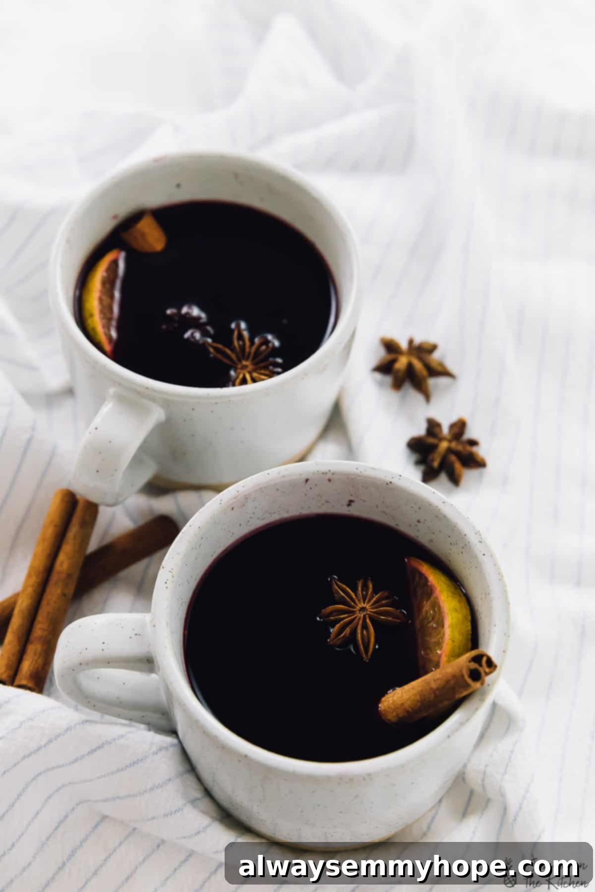 Two white mugs brimming with aromatic spiced mulled wine, garnished with orange slices and star anise, resting on a striped fabric, evoking warmth and holiday cheer.