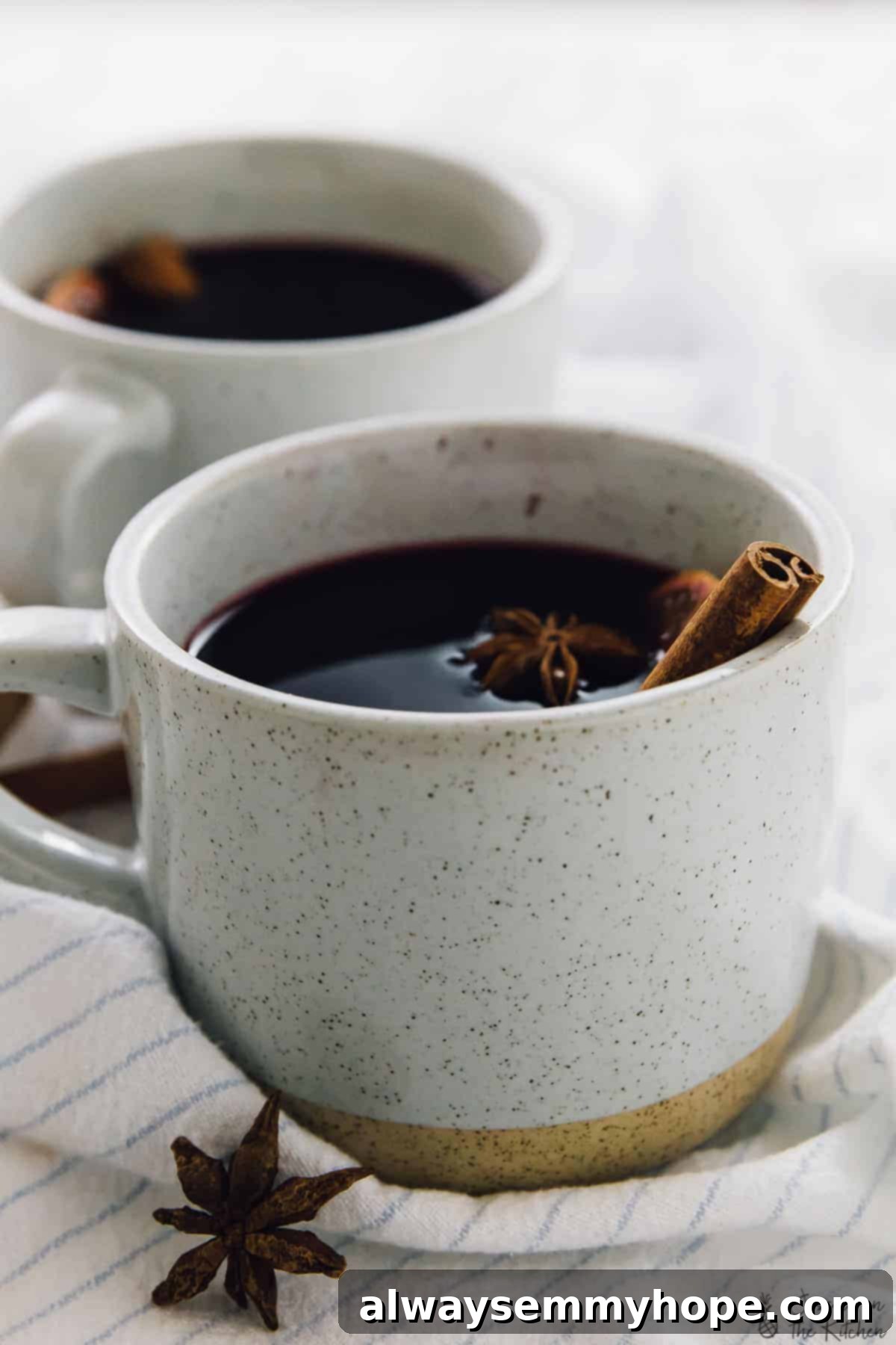 Two white mugs of spiced mulled wine, elegantly garnished with fresh orange slices and star anise, creating a warm and inviting display on a striped kitchen towel.
