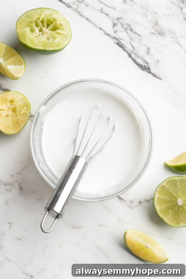 Overhead view of a small bowl of creamy vegan lime crema with a whisk, showing its smooth texture and fresh green lime zest.