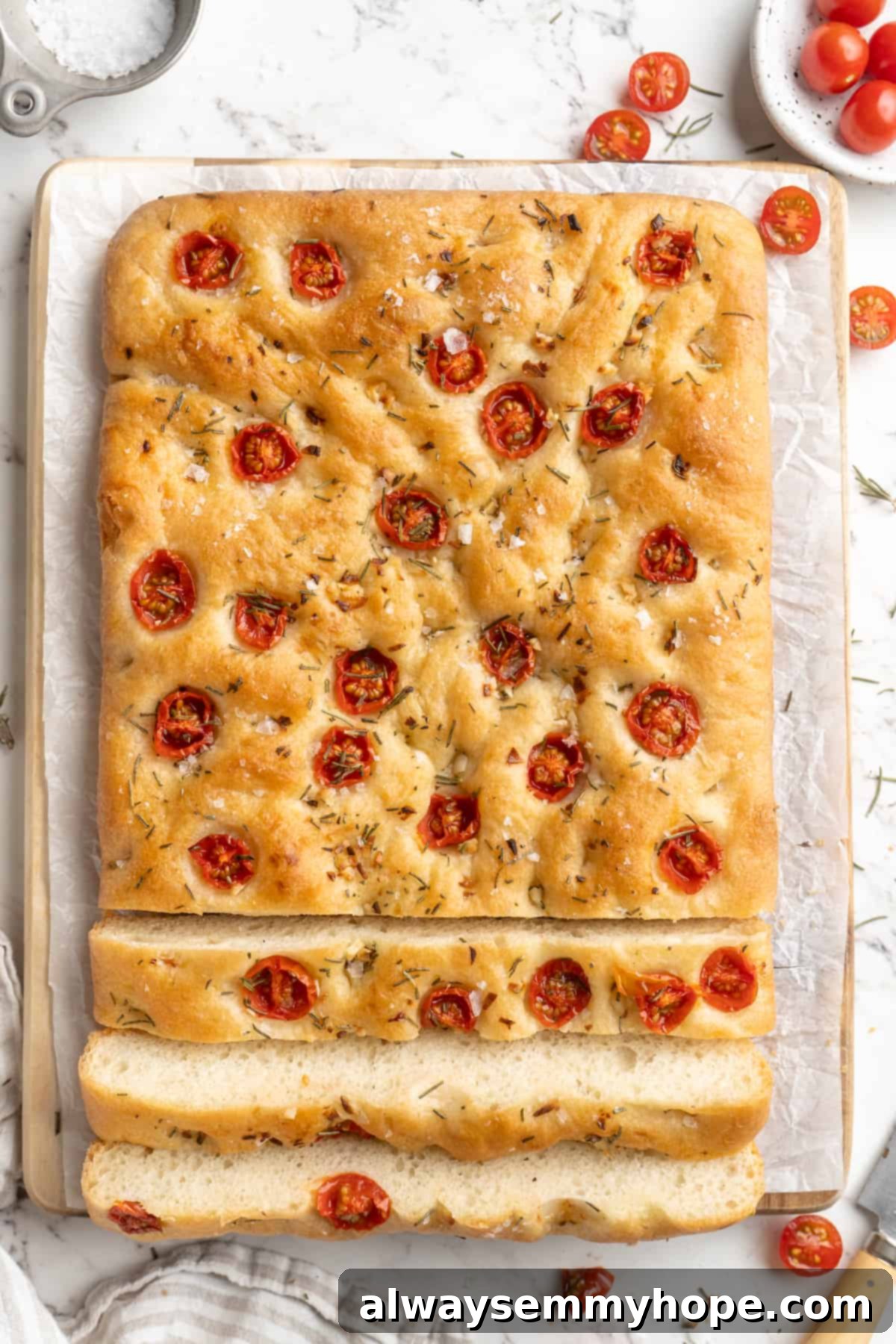 Half-sliced gluten-free focaccia loaf revealing its soft interior, topped with tomatoes and rosemary, ready to be served.