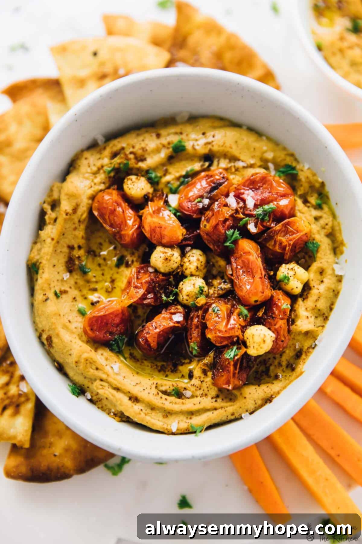 This roasted red pepper hummus recipe is sweet, savoury, smoky, and spicy. It's easy to make and ready to impress! Overhead view of spicy roasted red pepper hummus in a white bowl with carrots and toasted pita chips on the side.