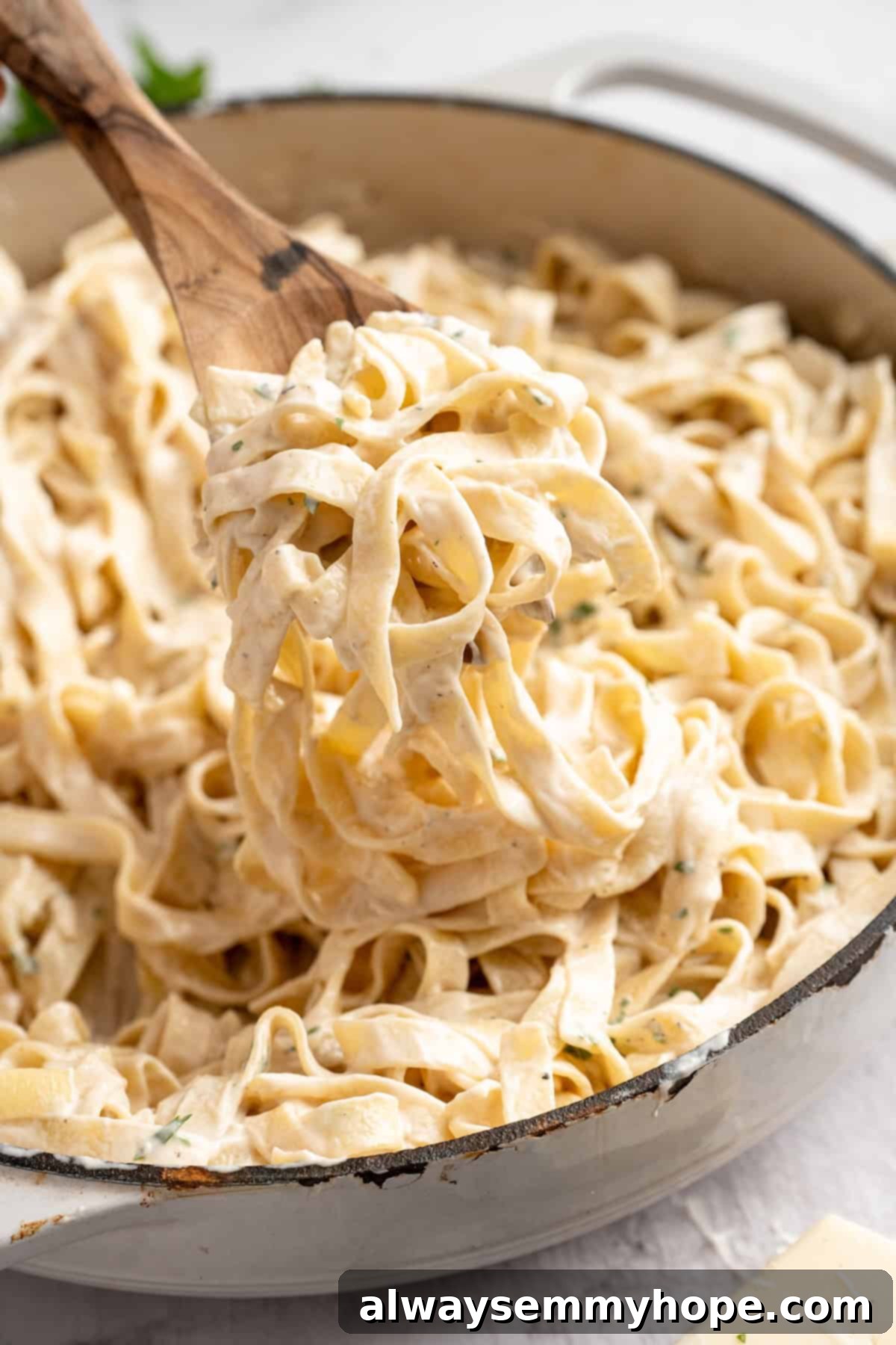 Lifting a portion of vegan garlic Alfredo pasta from a skillet with a wooden spoon, showing its creamy texture.