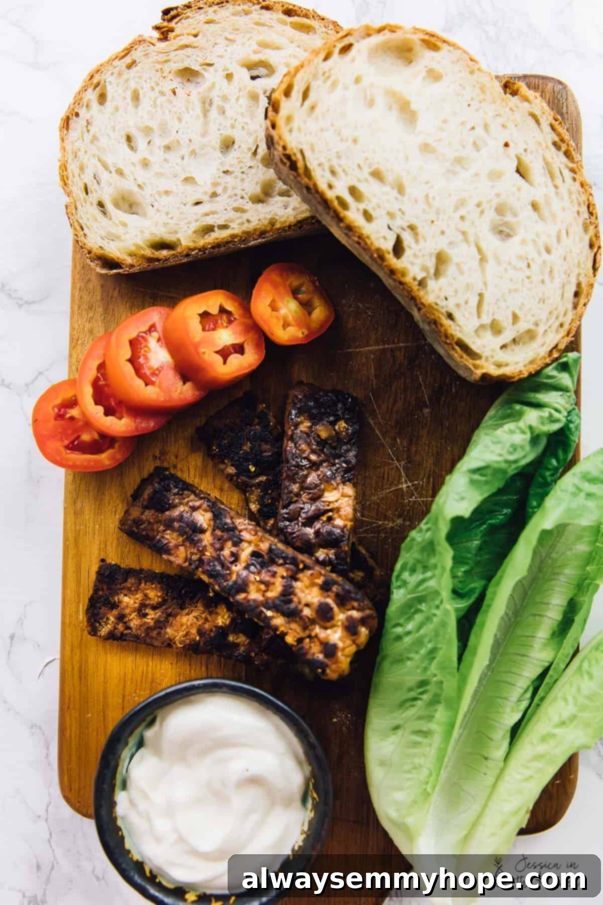 Fresh ingredients for a vegan BLT sandwich, including tempeh, lettuce, tomato, bread, and spices, arranged for a bird's eye view.