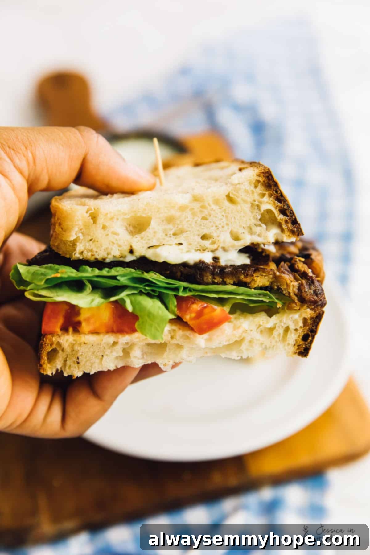 A person holding half of a delicious vegan BLT sandwich, highlighting the textures of the tempeh bacon, fresh lettuce, and tomato.
