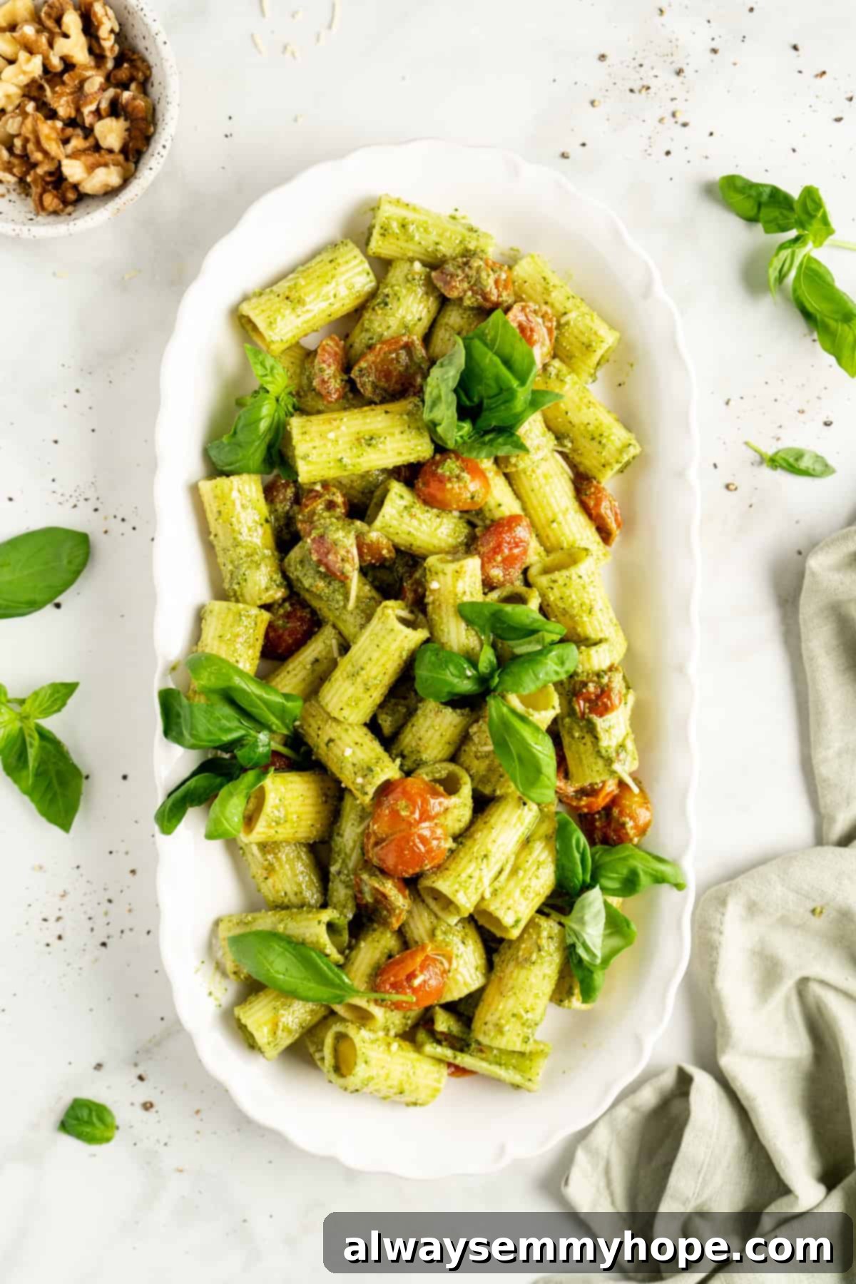 Overhead view of pesto pasta with basil and cherry tomatoes in white dish with small bowl of nuts.